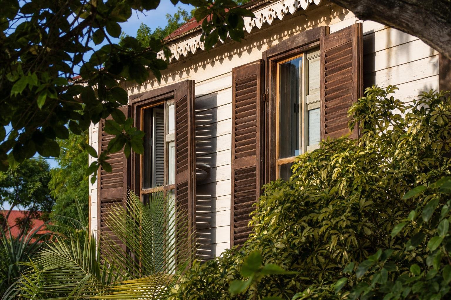 Façade en bois d'une maison créole aux persiennes entrouvertes, entourée de végétation